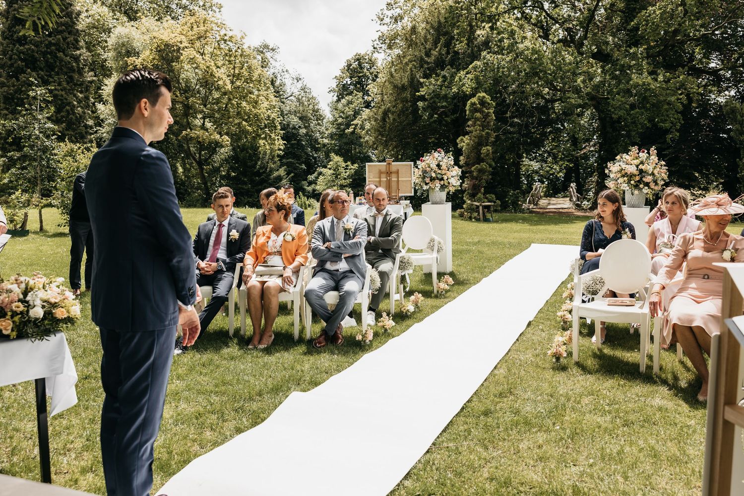 Groom in navy suit stands at outdoor wedding aisle awaiting bride, guests seated on white chairs in sunny garden.