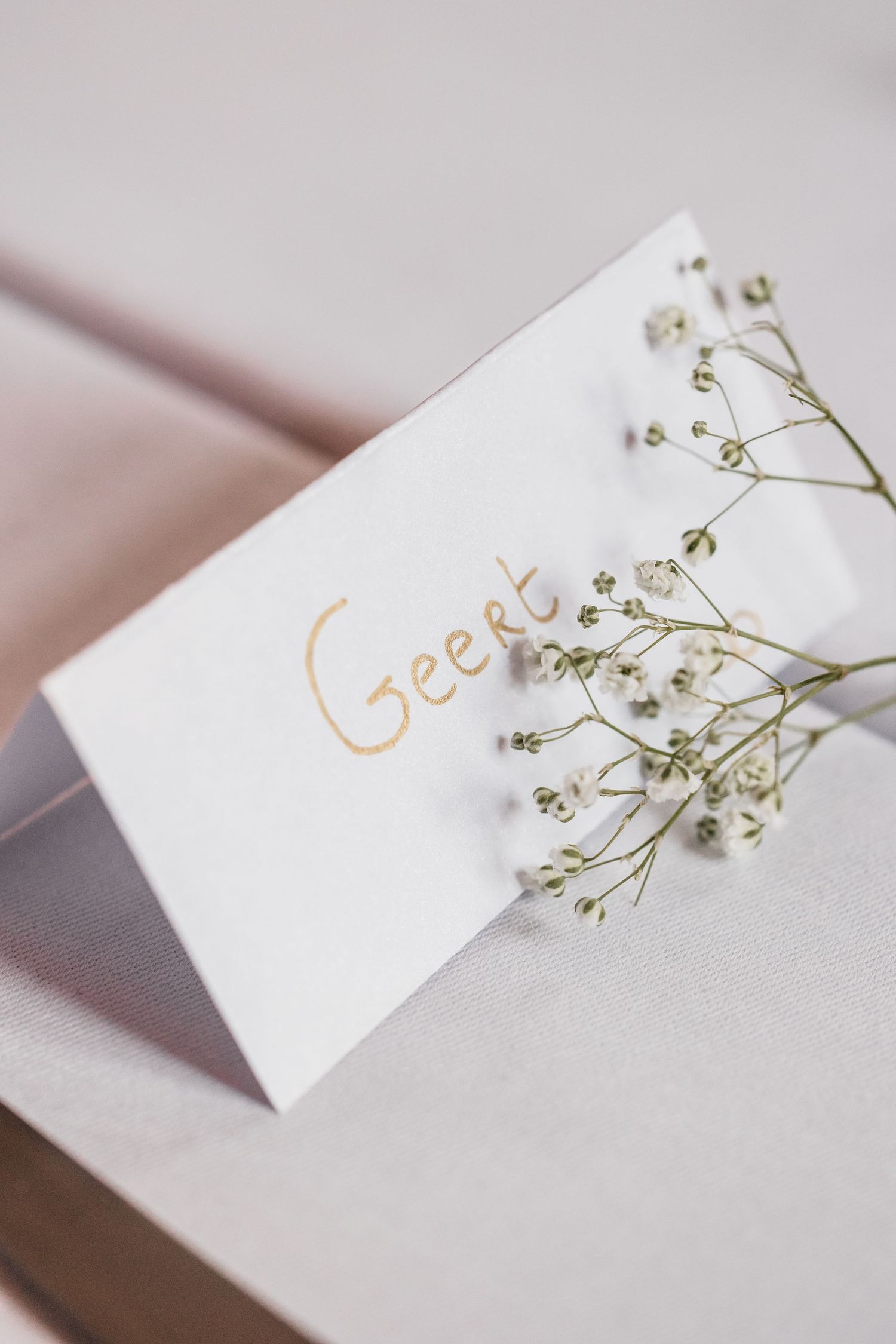 Close-up of elegant white wedding place card with gold handwritten name Geert and delicate baby's breath flowers.