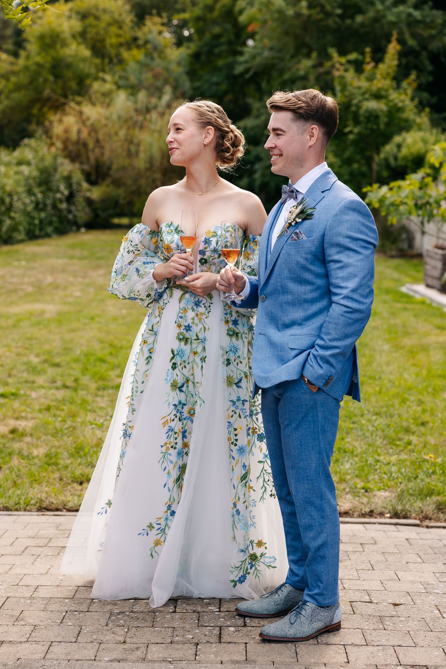 Bride in floral gown and groom in blue suit holding champagne in garden.