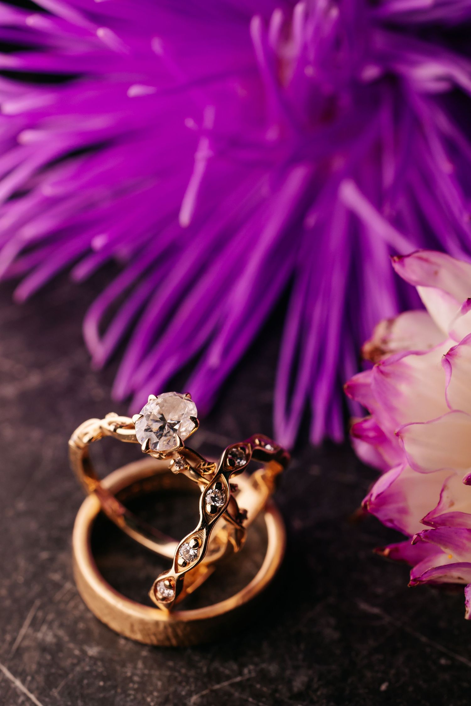 Two ornate gold wedding rings with diamonds displayed beside vibrant purple and pink flowers on dark surface.