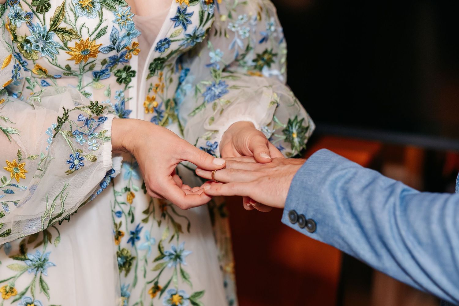 Groom in blue suit places wedding ring on brides finger during indoor wedding ceremony.