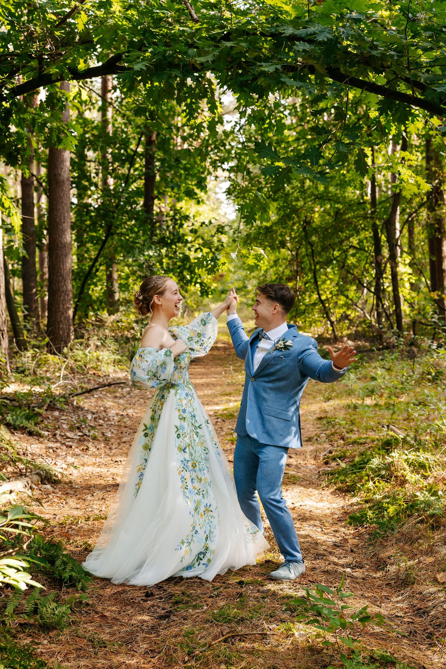 Bride and groom dancing joyfully on a forest path, bride wearing a floral wedding dress, groom in a blue suit.