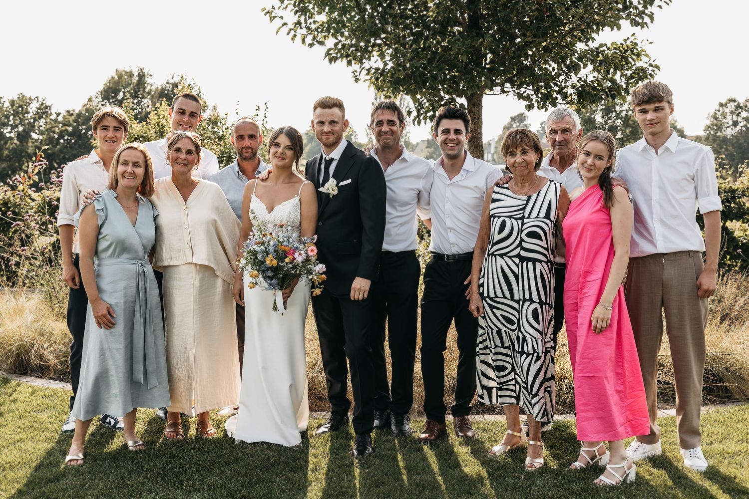 Large wedding family group photo outdoors under a tree, featuring bride in white gown holding a colorful bouquet.