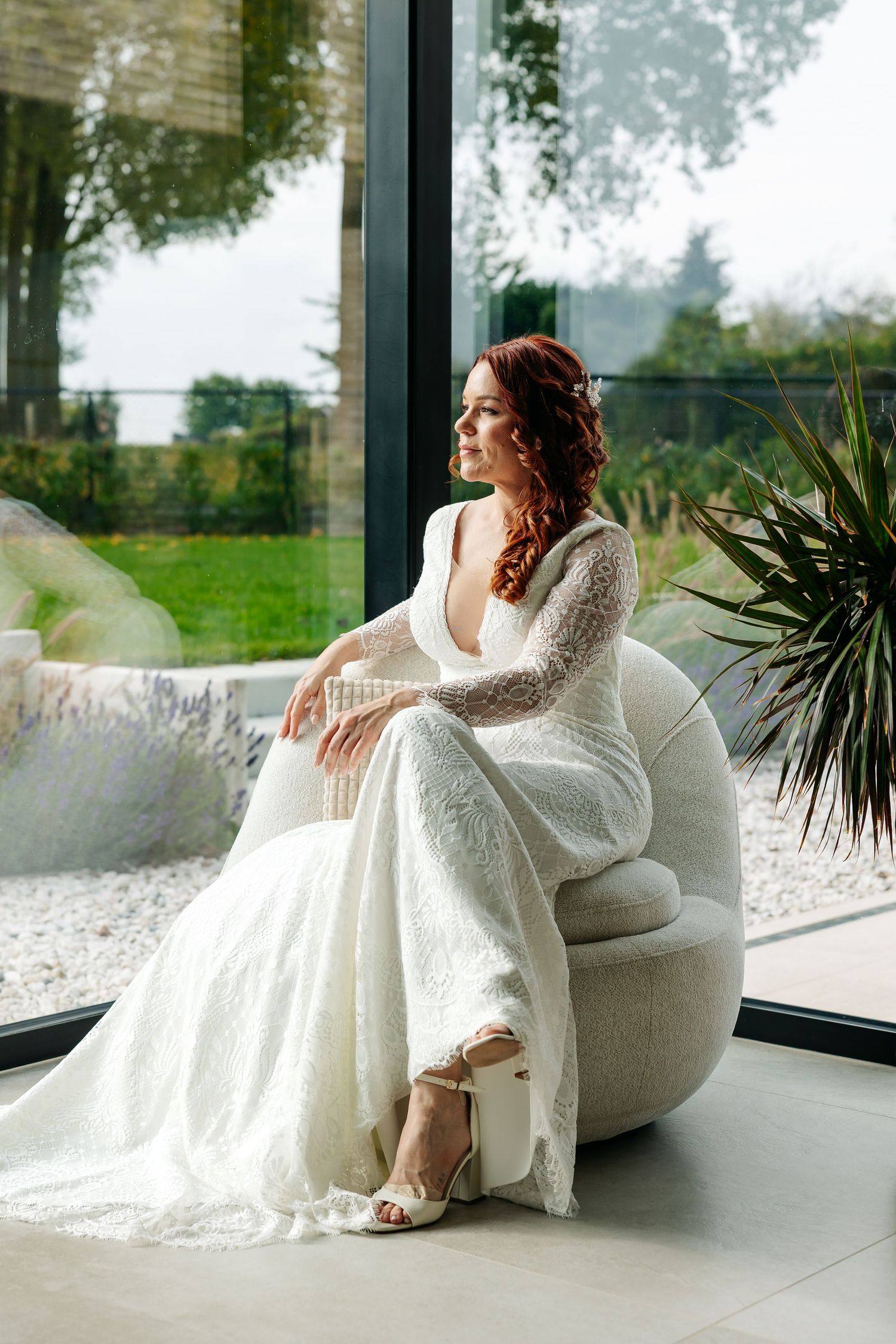 Stunning red-haired bride in deep V-back lace gown sits in modern chair by large glass windows.