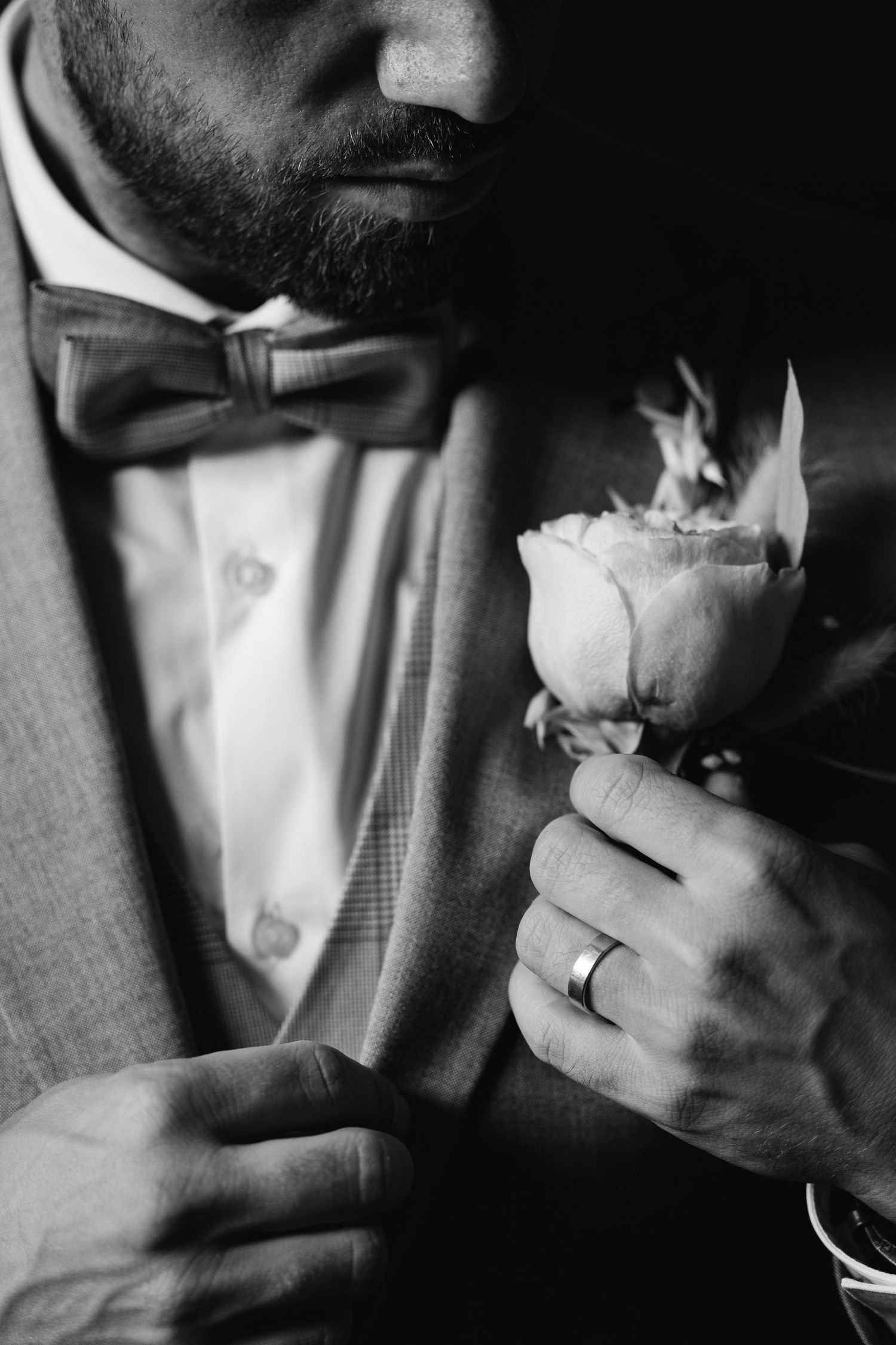 Black and white close-up of a bearded groom in a suit holding a rose boutonniere with a wedding ring.