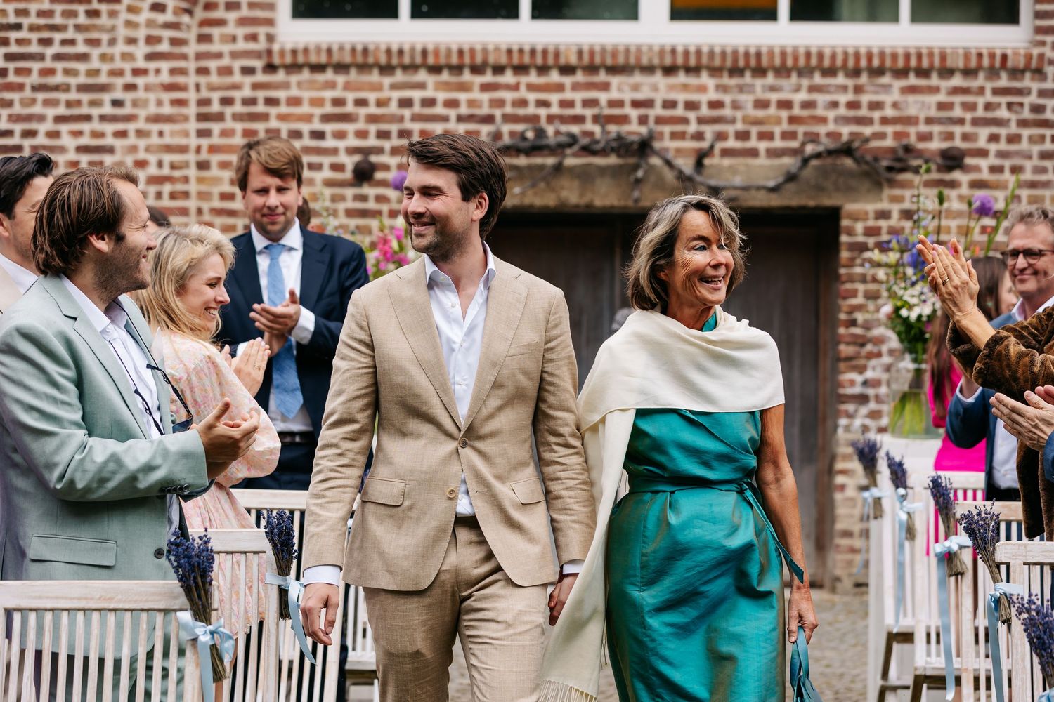 A man in a beige suit and woman in teal walk through applauding guests at an outdoor wedding venue.