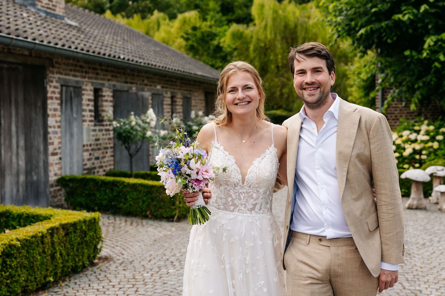 Smiling bride holding colorful bouquet stands with groom in tan suit outside rustic stone venue.