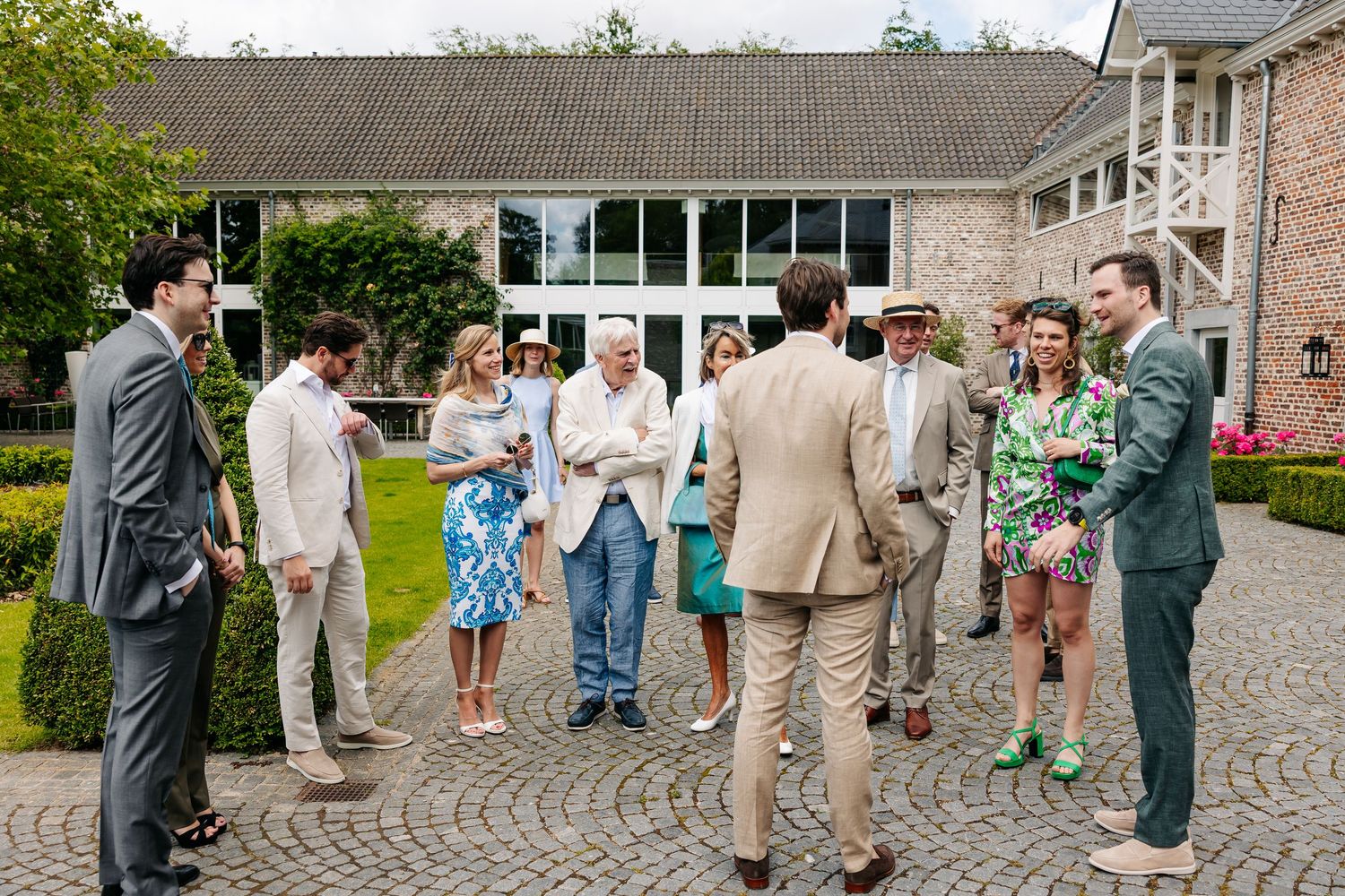 Wedding guests in elegant summer attire socializing on a cobblestone courtyard outside a rustic stone venue.