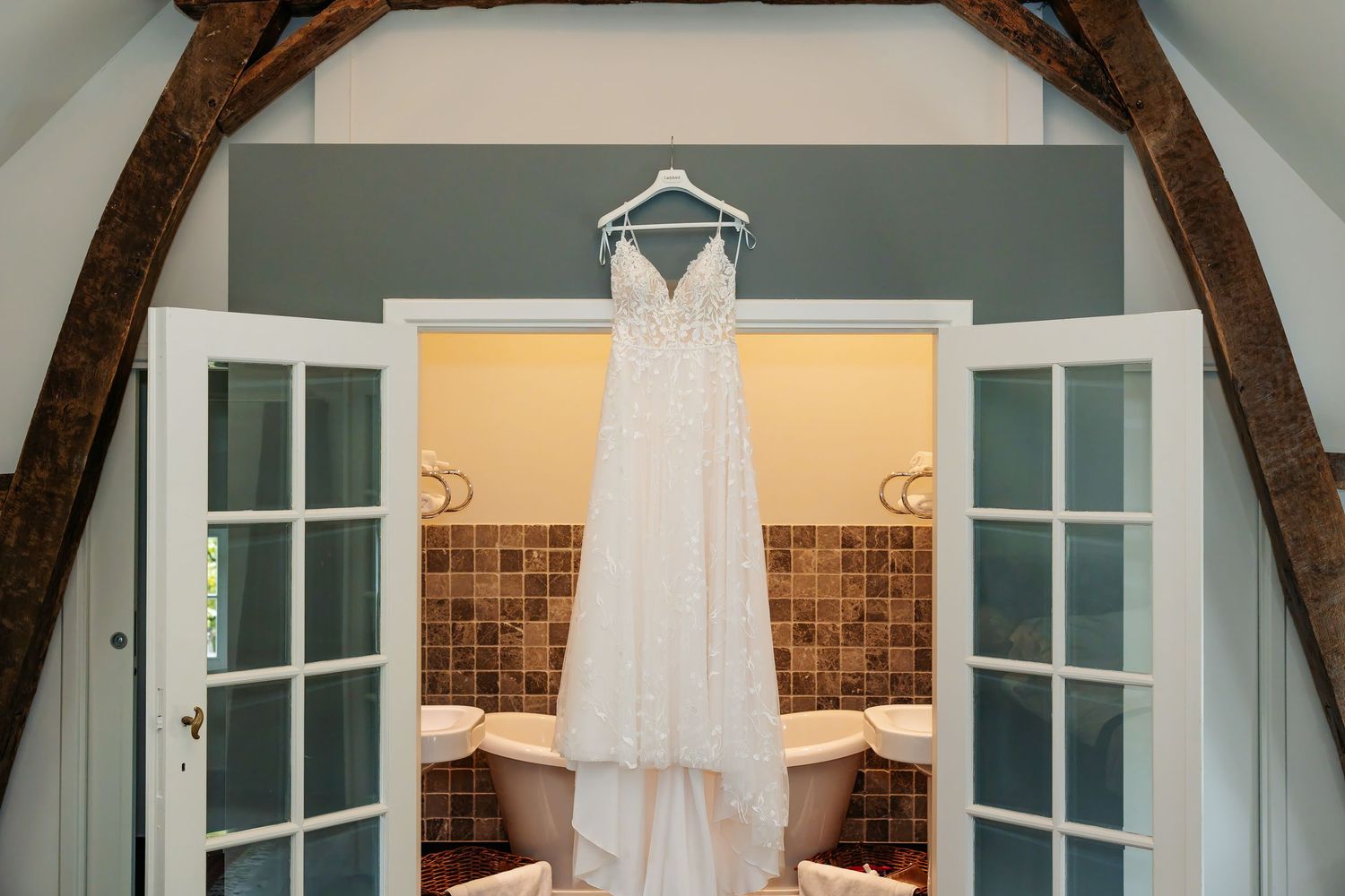 White lace wedding dress hanging on French doors above a freestanding bathtub in rustic venue.