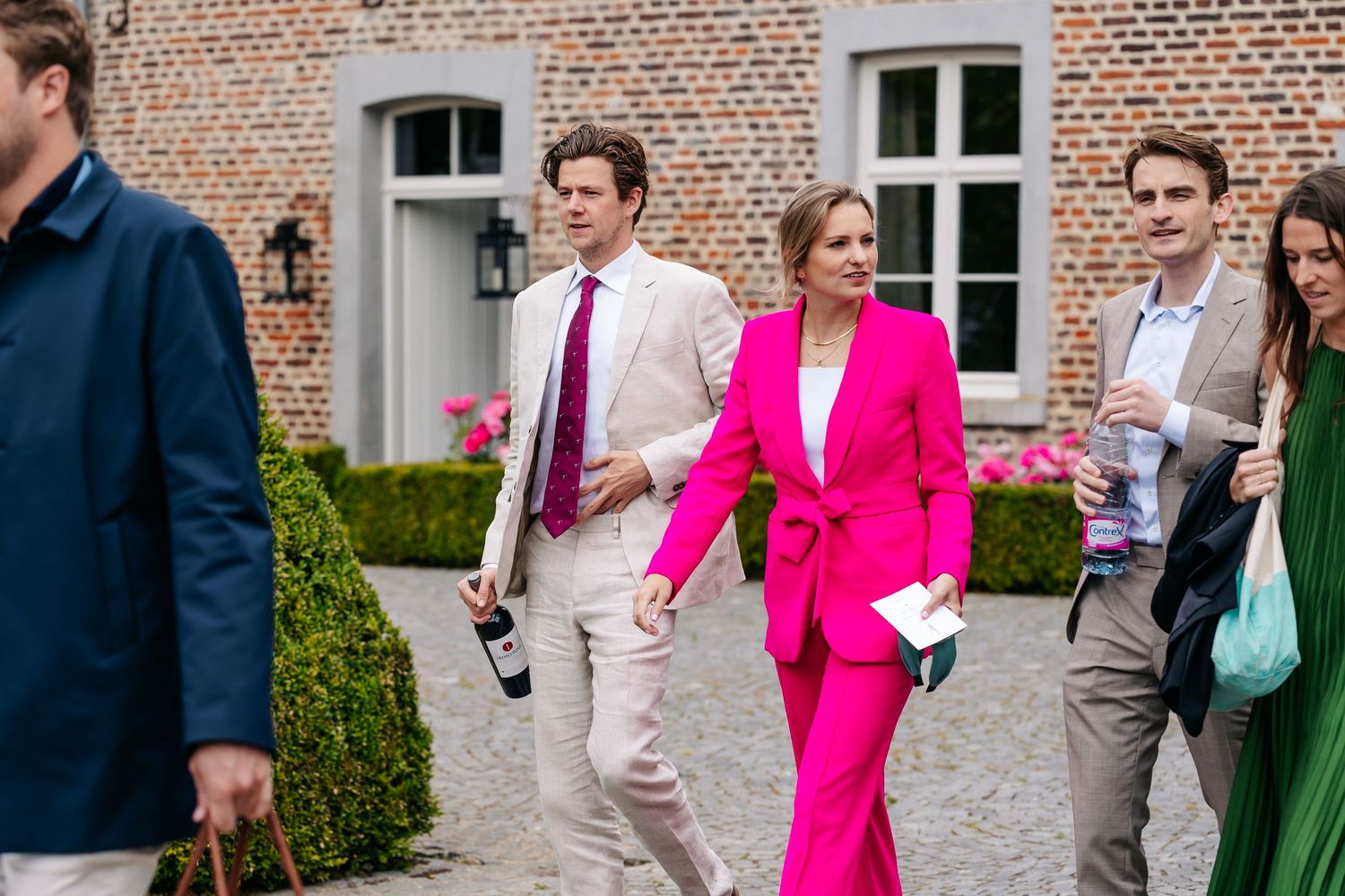 Wedding guests walking outdoors, woman in bright pink suit and man in white suit leading the group at a brick estate.