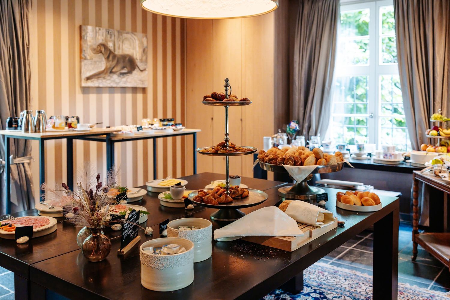 Elegant hotel breakfast buffet spread with pastries, breads, and assorted foods on a dark wood table in a warmly lit dining room.