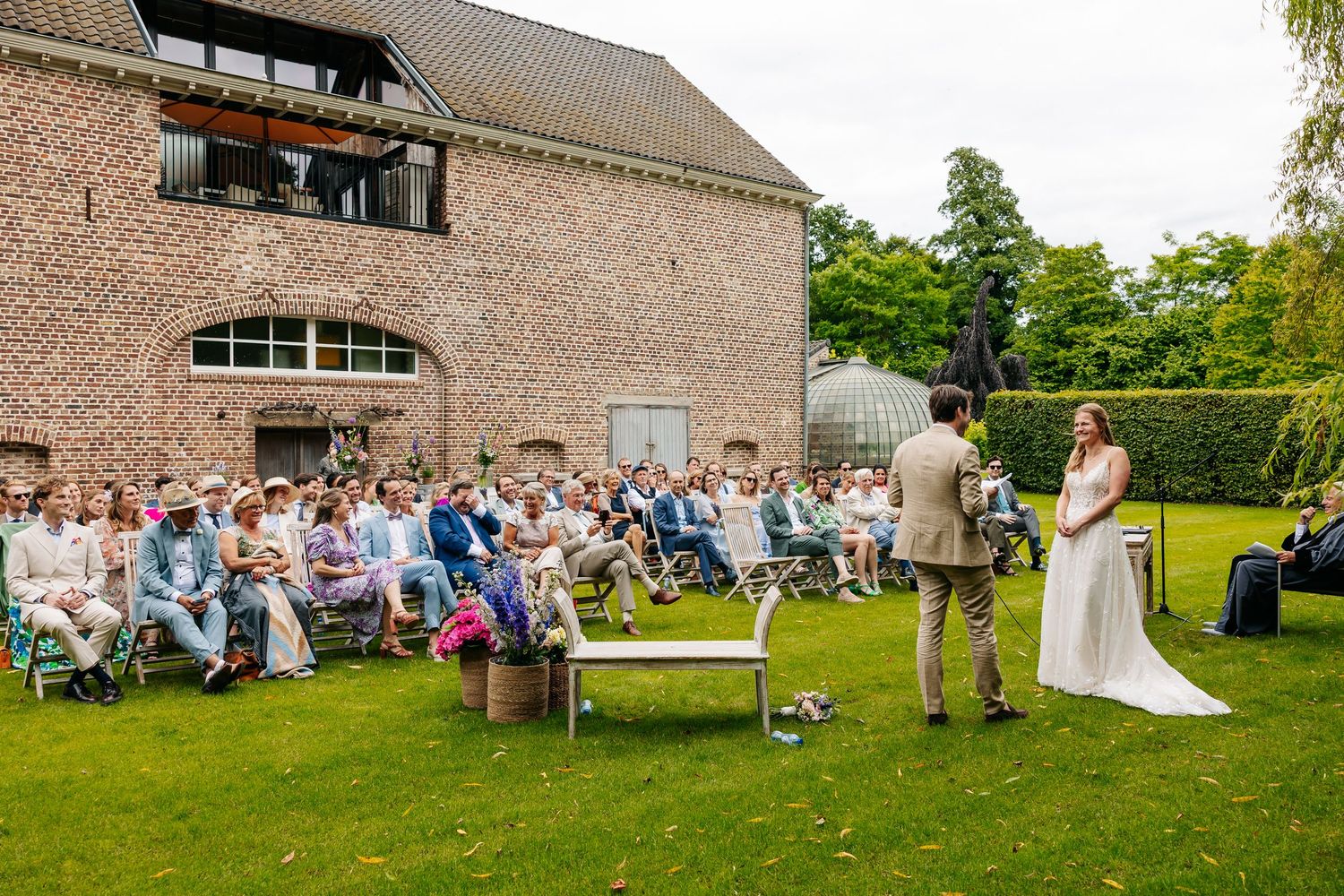 Outdoor wedding ceremony on green lawn with guests seated before brick building and willow tree.