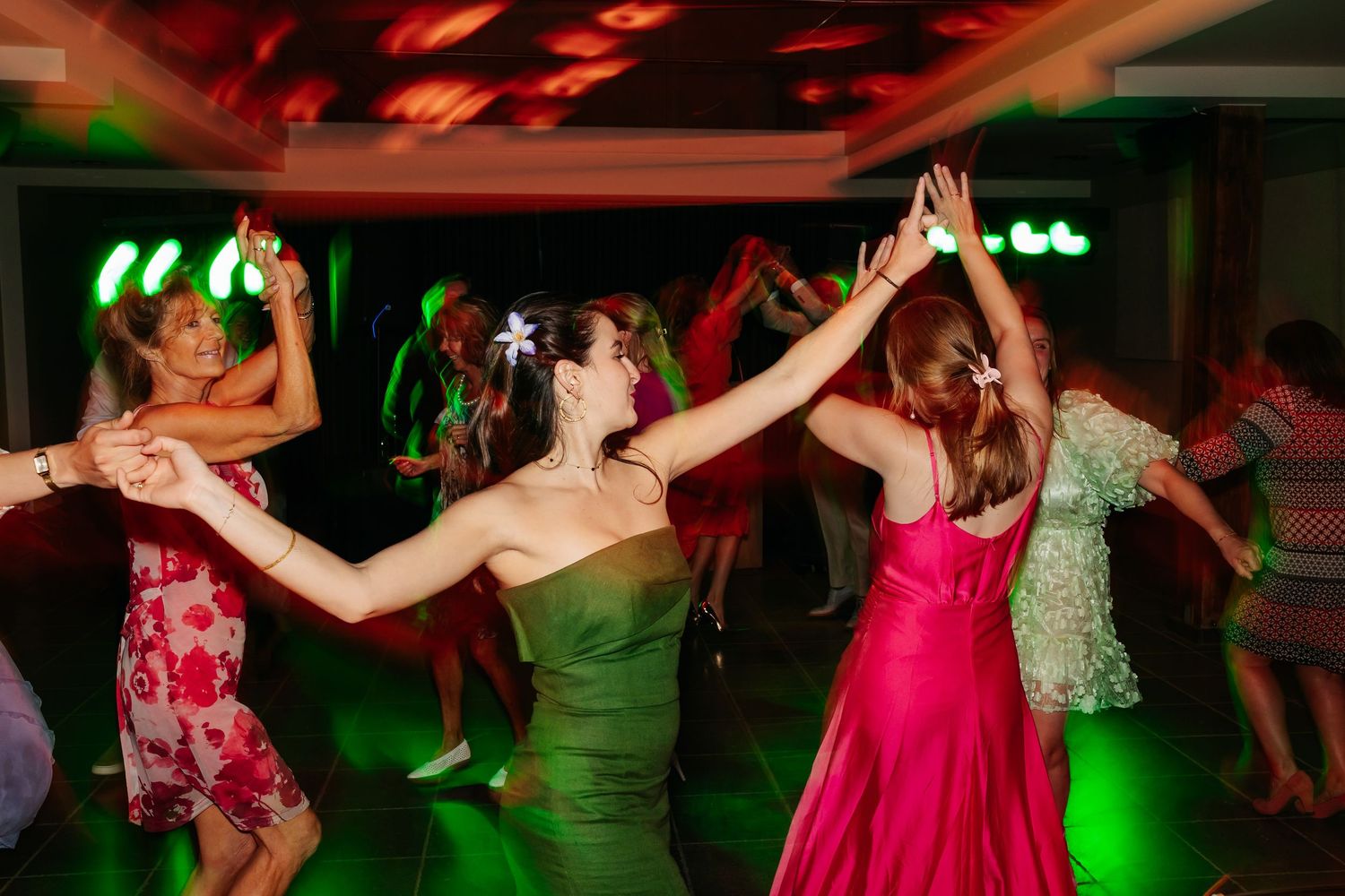 Wedding guests in colorful dresses dancing together under green lights at reception.