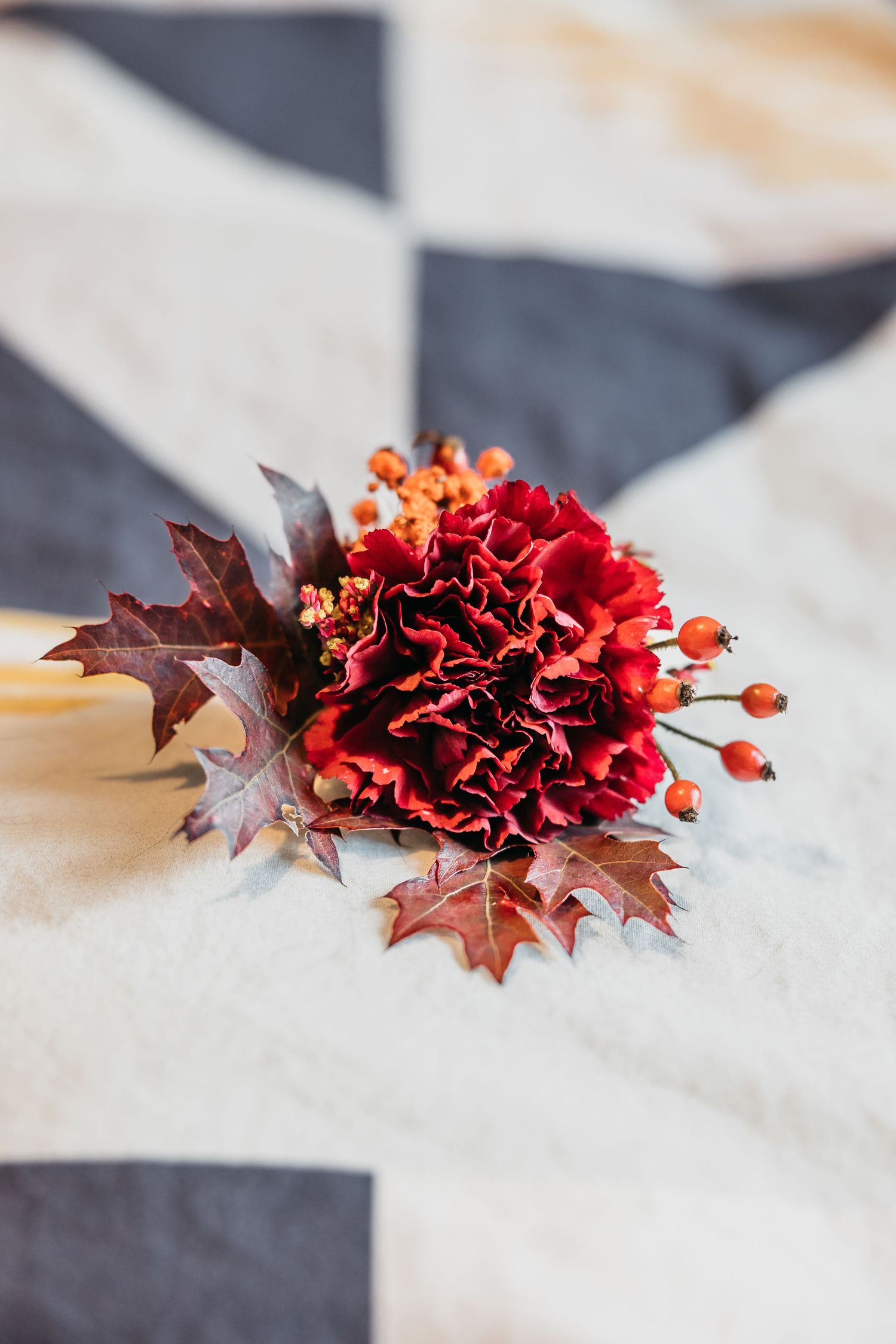 Elegant autumn wedding boutonniere with red carnation, berries, and maple leaves on fabric.