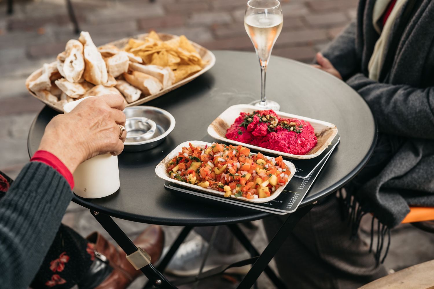 People enjoying outdoor dining with colorful dishes and champagne on a round black table.