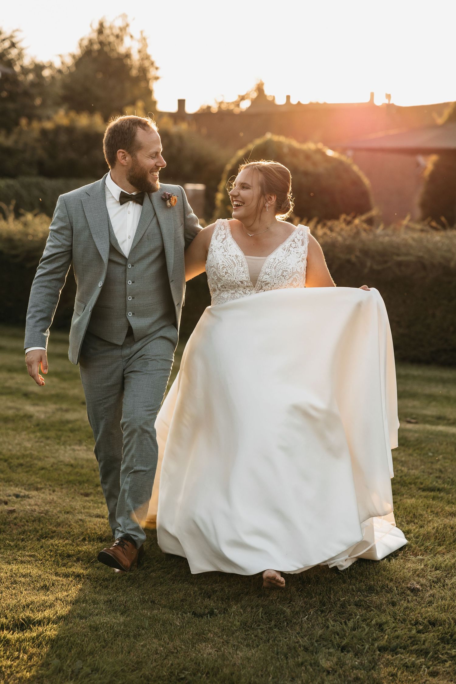 Laughing bride and groom walking together across garden lawn in warm golden sunset.