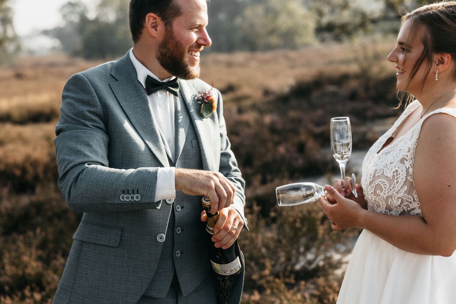 Groom in gray suit opening champagne bottle for bride in white lace wedding dress outdoors.