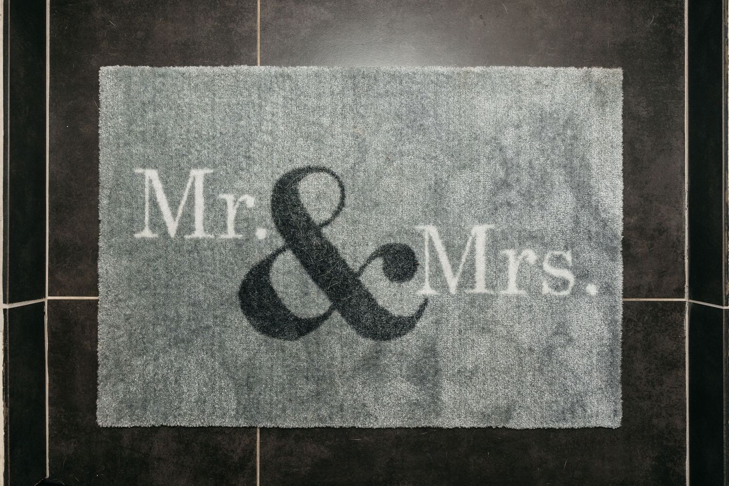 Gray bath mat with Mr. & Mrs. text in white and black lettering on dark tile floor.
