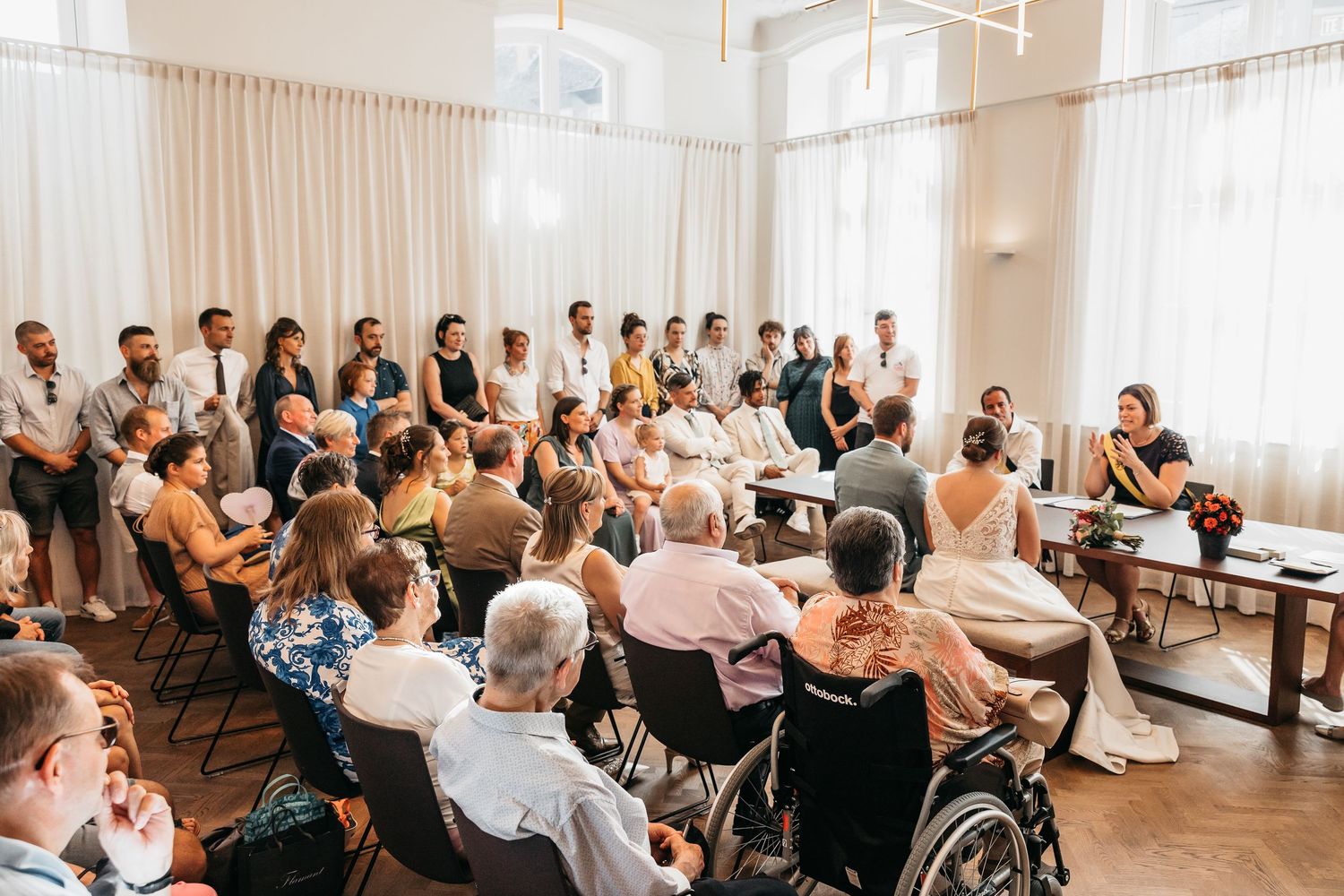 Wide shot of wedding ceremony with guests seated and standing in bright white decorated hall.