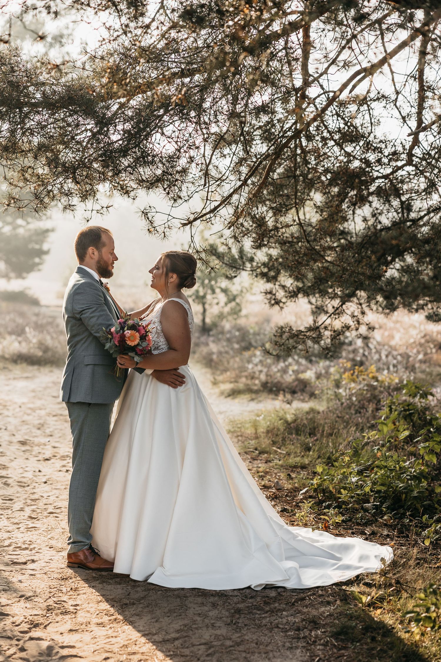 Bride and groom embrace under pine trees on a sandy forest path.