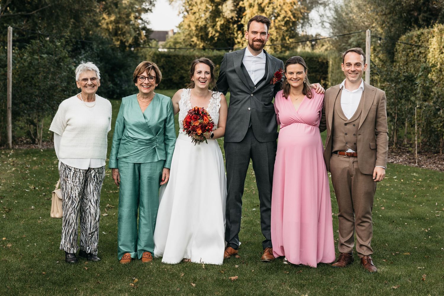 Bride and groom with four family members smiling in garden at outdoor wedding with red bridal bouquet.