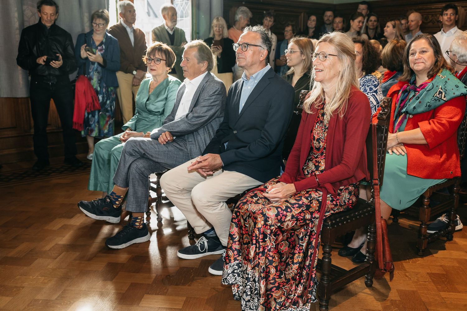 Rows of smiling wedding guests in colorful formal attire seated in a grand wood-paneled ceremony hall.