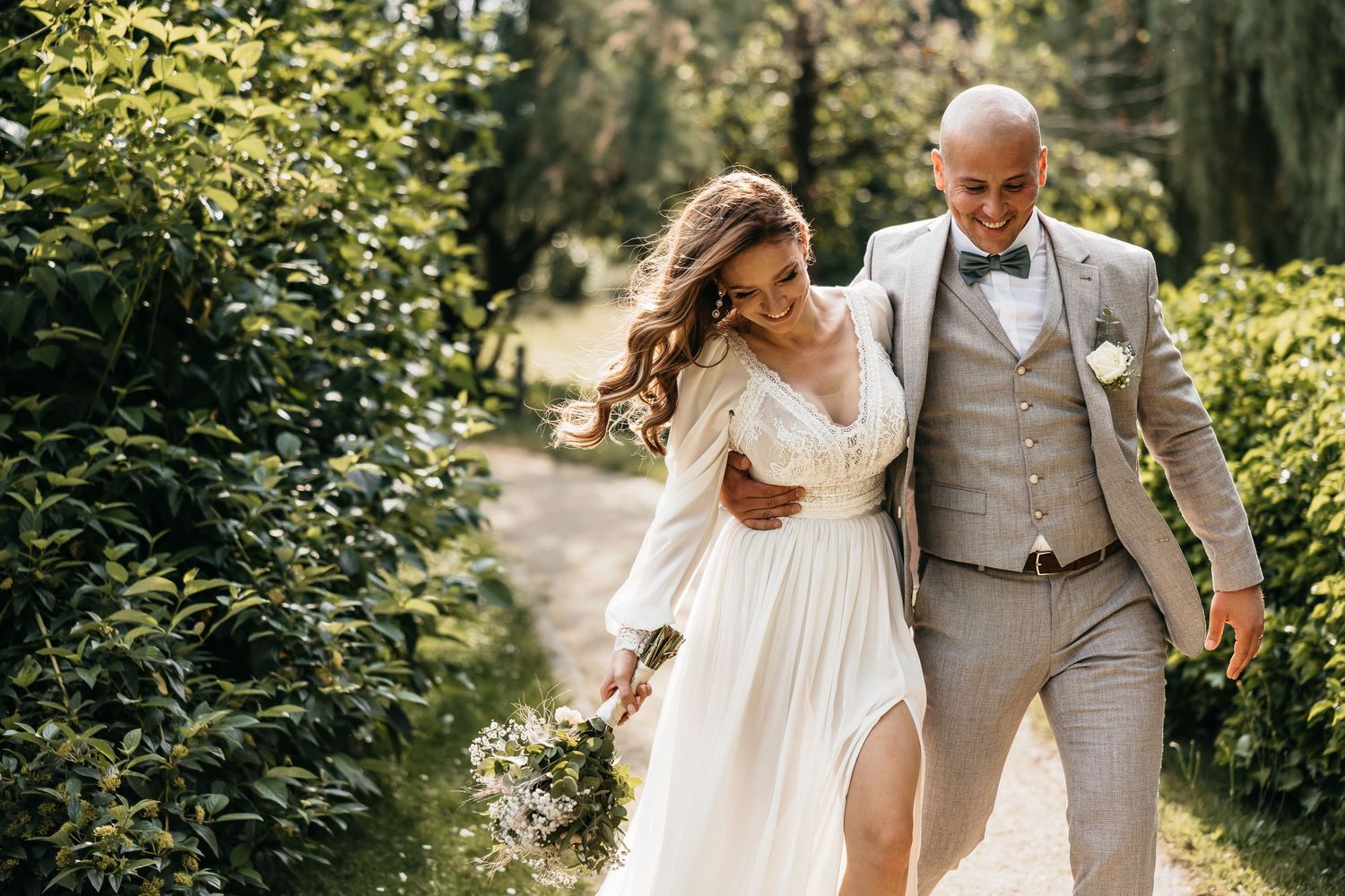 Smiling bride and groom walking arm in arm along a sun-dappled garden path on their wedding day.