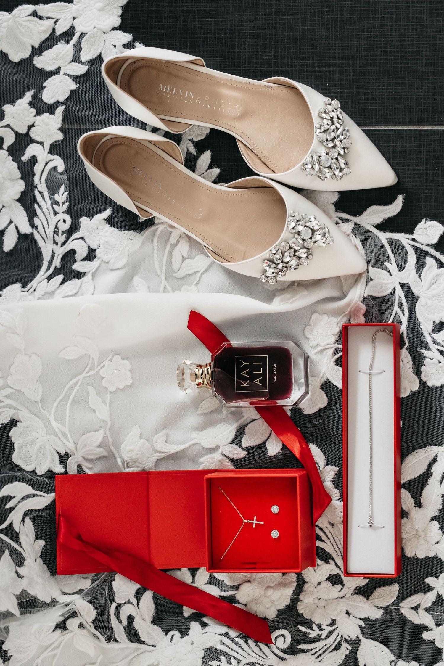 Bridal flat lay with white jeweled shoes, Kayali perfume, red gift boxes with jewelry.