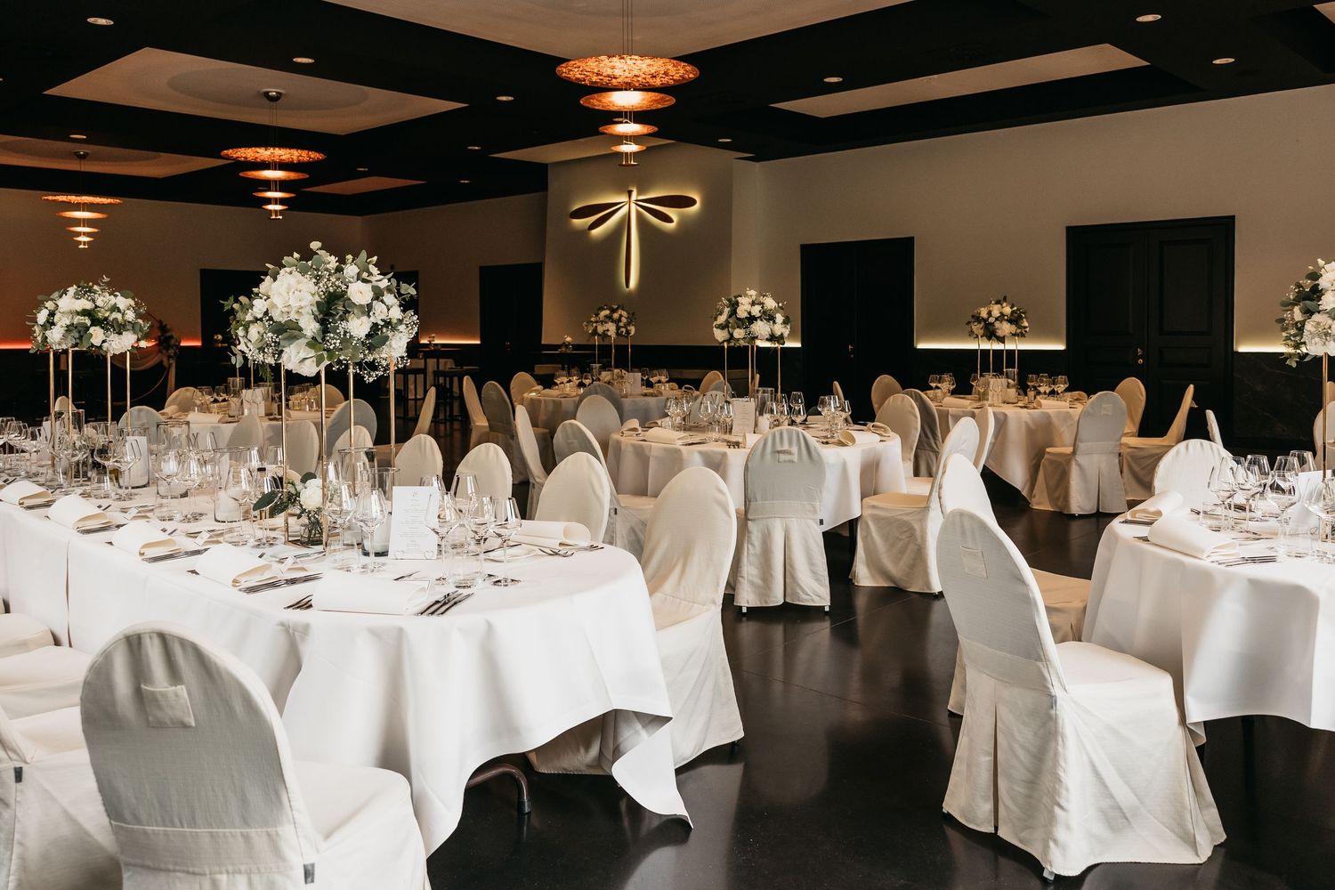 Elegant wedding reception hall with white chair covers, round tables, floral centerpieces, and warm pendant lighting.