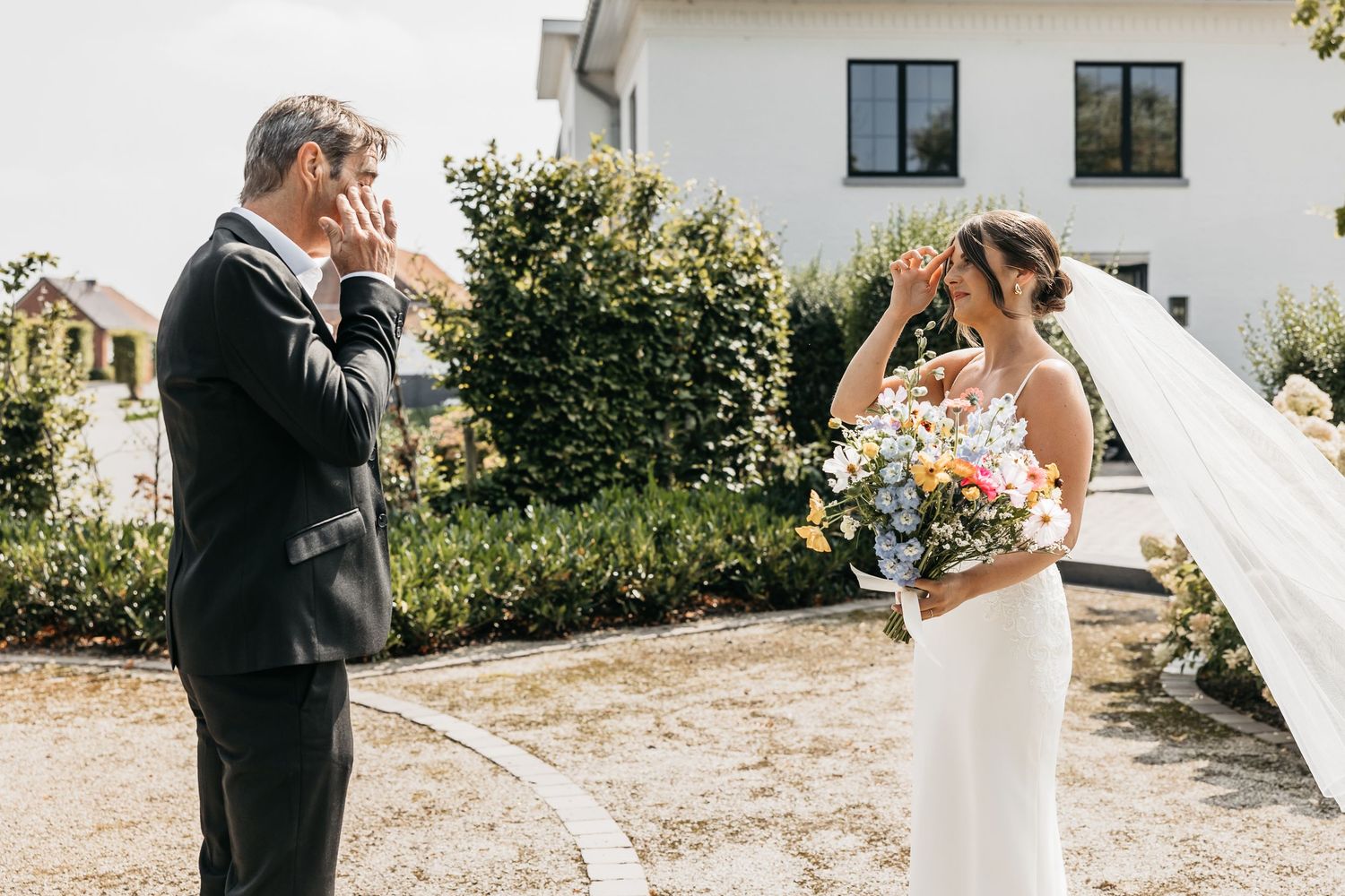 Bride and groom sharing emotional first look moment outdoors, both wiping tears of joy on wedding day.