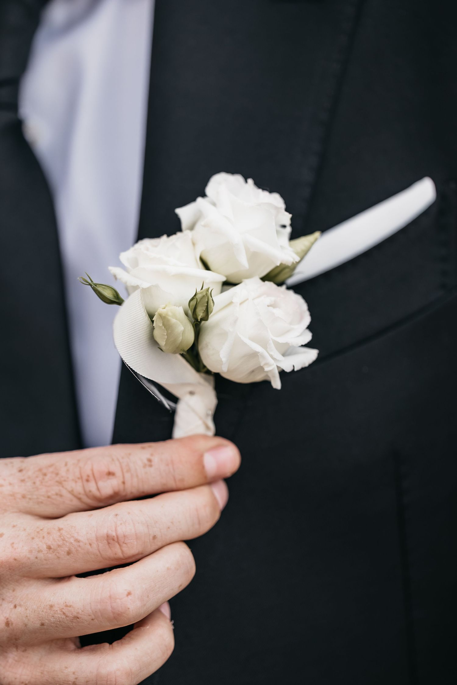 Close-up of groom holding delicate white rose boutonniere against dark wedding suit lapel.