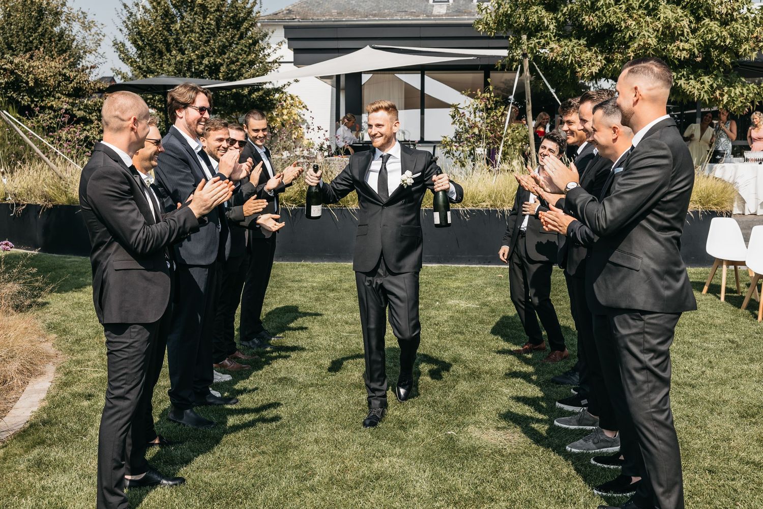 Groom walking through a tunnel of applauding groomsmen in black suits at outdoor wedding.