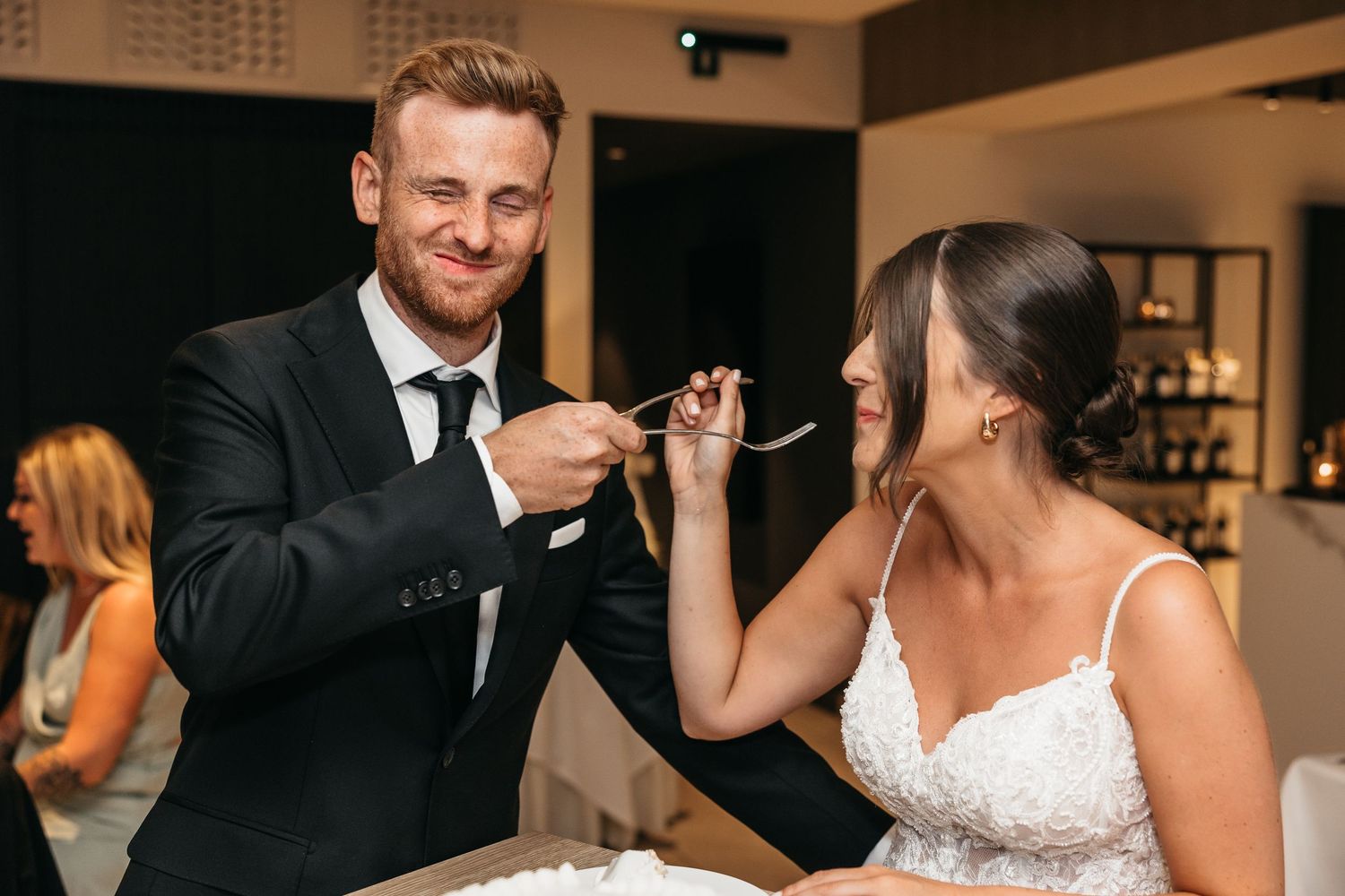 Smiling groom feeding bride a taste of wedding cake at elegant indoor reception dinner.