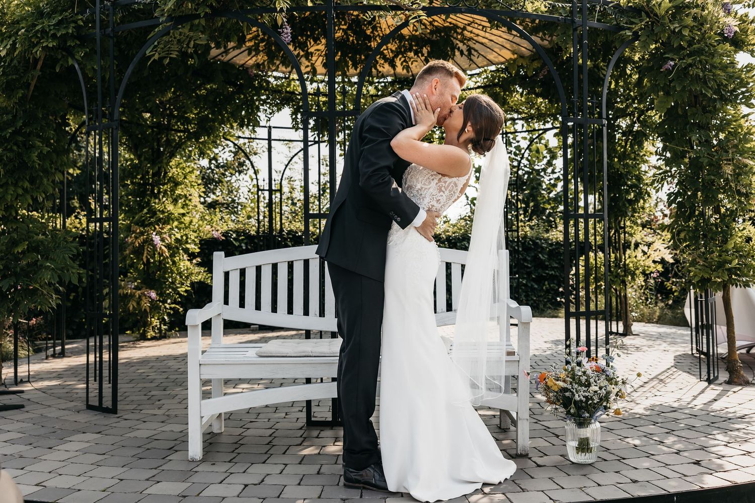 Bride and groom share passionate first kiss beside white bench under garden arbor.