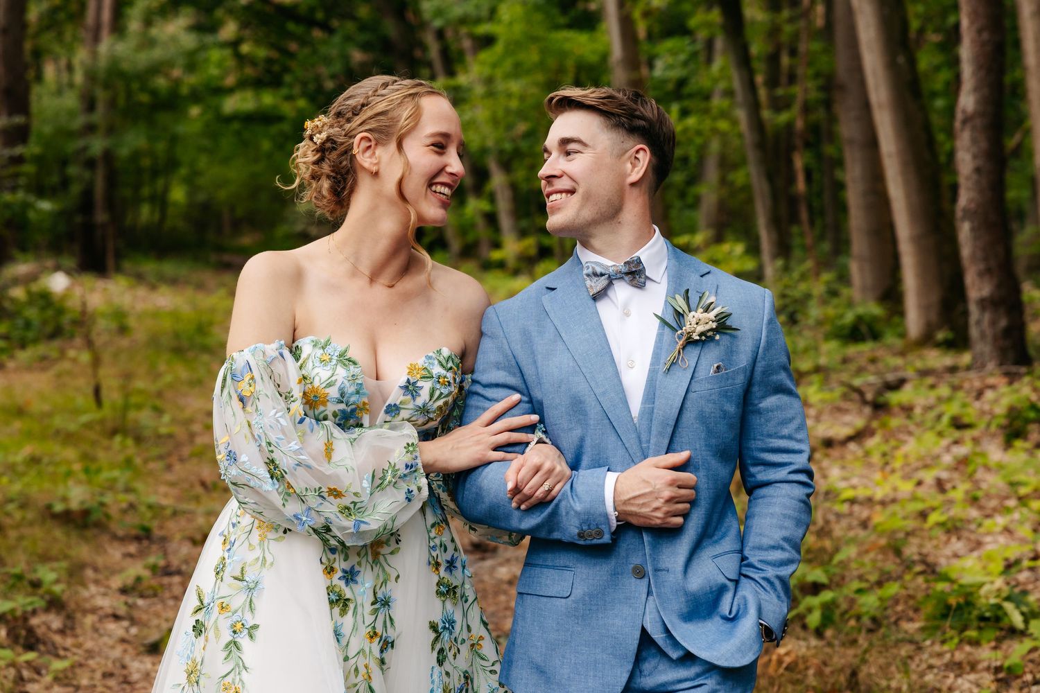 Bride and groom laughing together in forest wearing blue suit and floral dress.