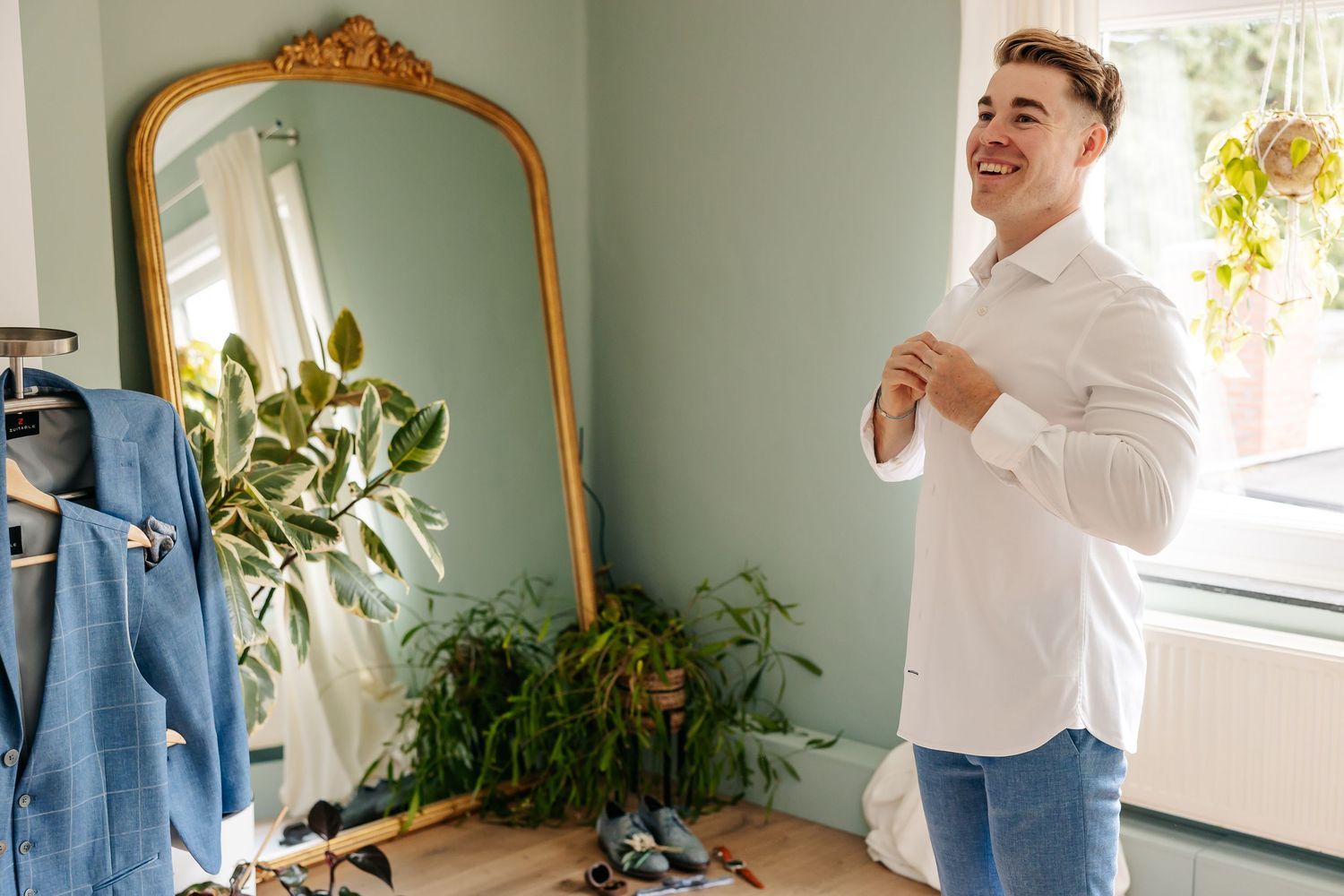 Smiling groom in white shirt buttoning up while standing in front of gold ornate mirror with green plants.