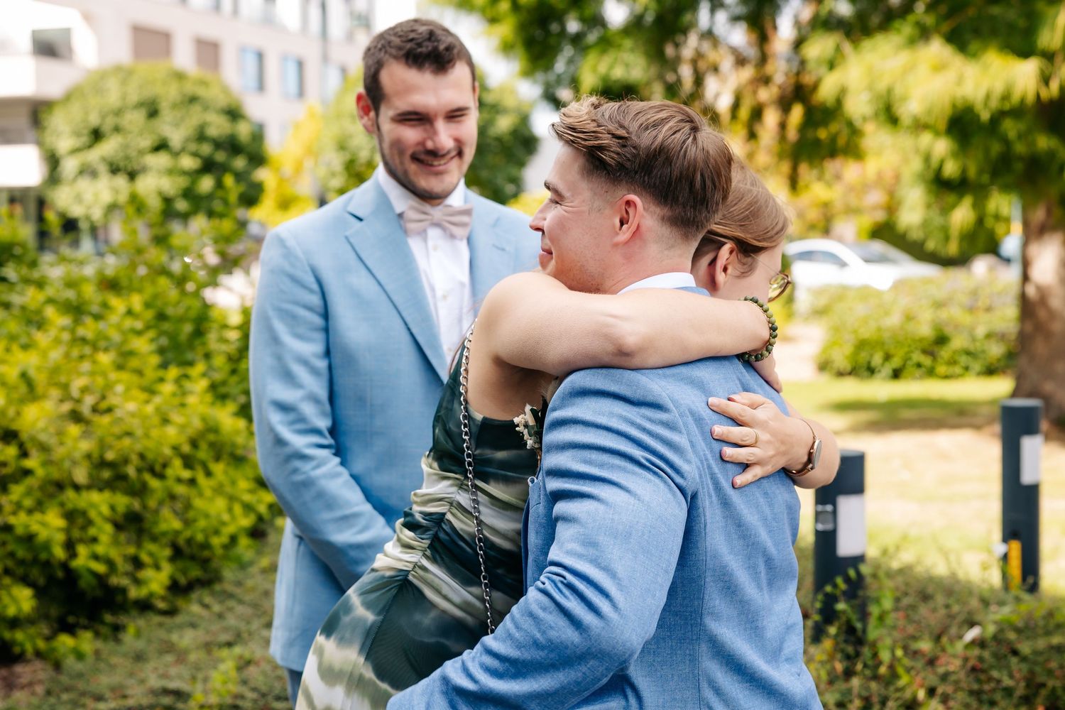 A man in a light blue suit smiles as two guests embrace at an outdoor wedding gathering.