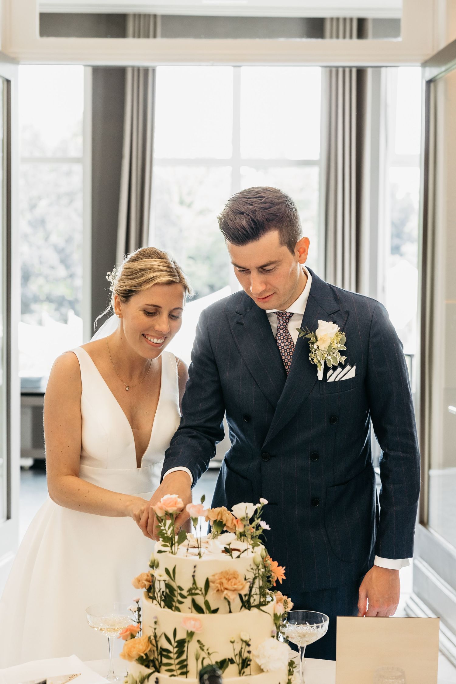 Bride and groom smiling while cutting their floral-decorated white wedding cake together indoors.