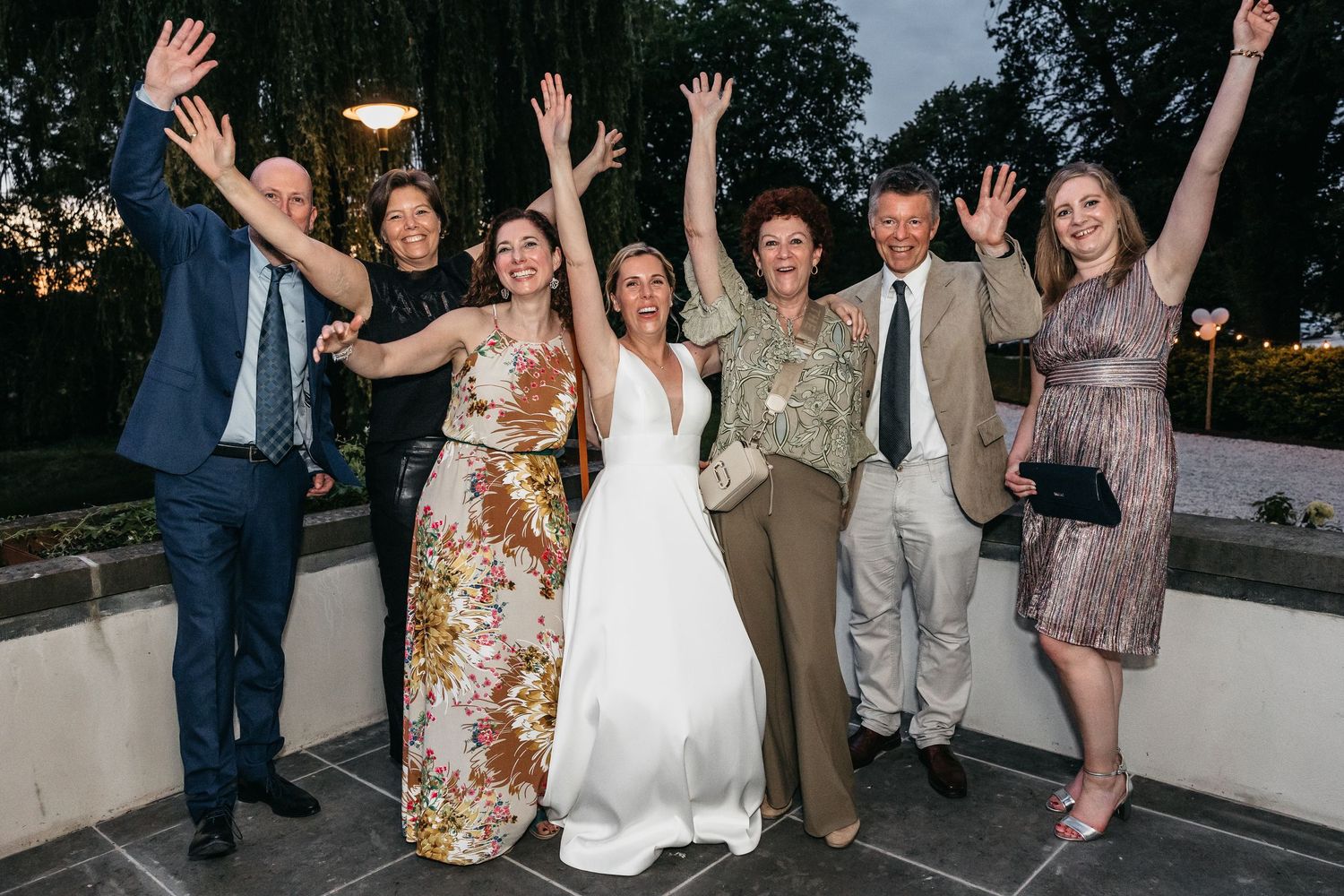 Bride in white gown celebrates with group of smiling guests with raised arms at wedding.