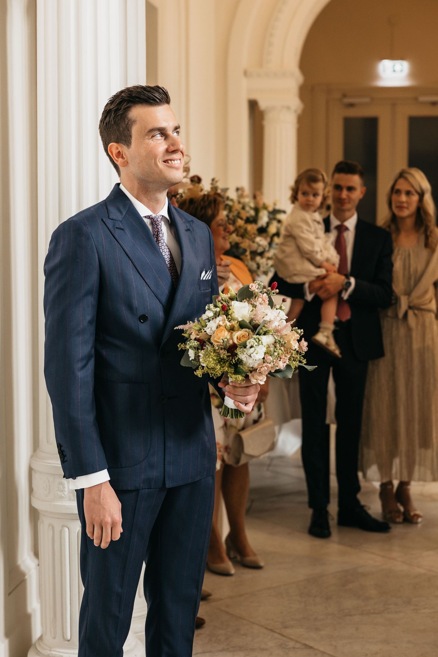 Smiling groom in navy suit holds wedding bouquet as guests gather behind him.