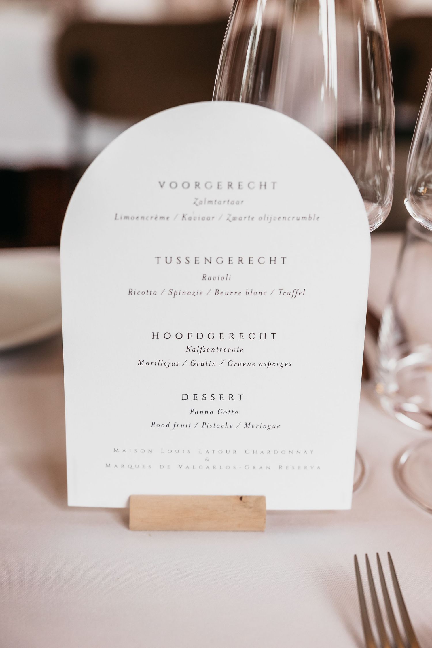 Elegant arch-shaped wedding menu card in Dutch displayed on a wooden stand beside crystal glasses.