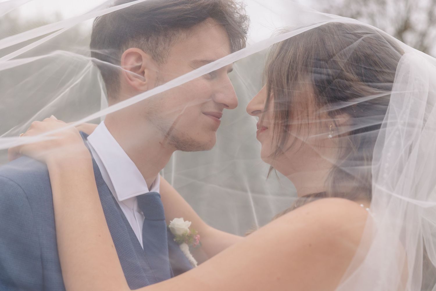 Bride and groom gazing at each other tenderly under a soft white wedding veil outdoors.