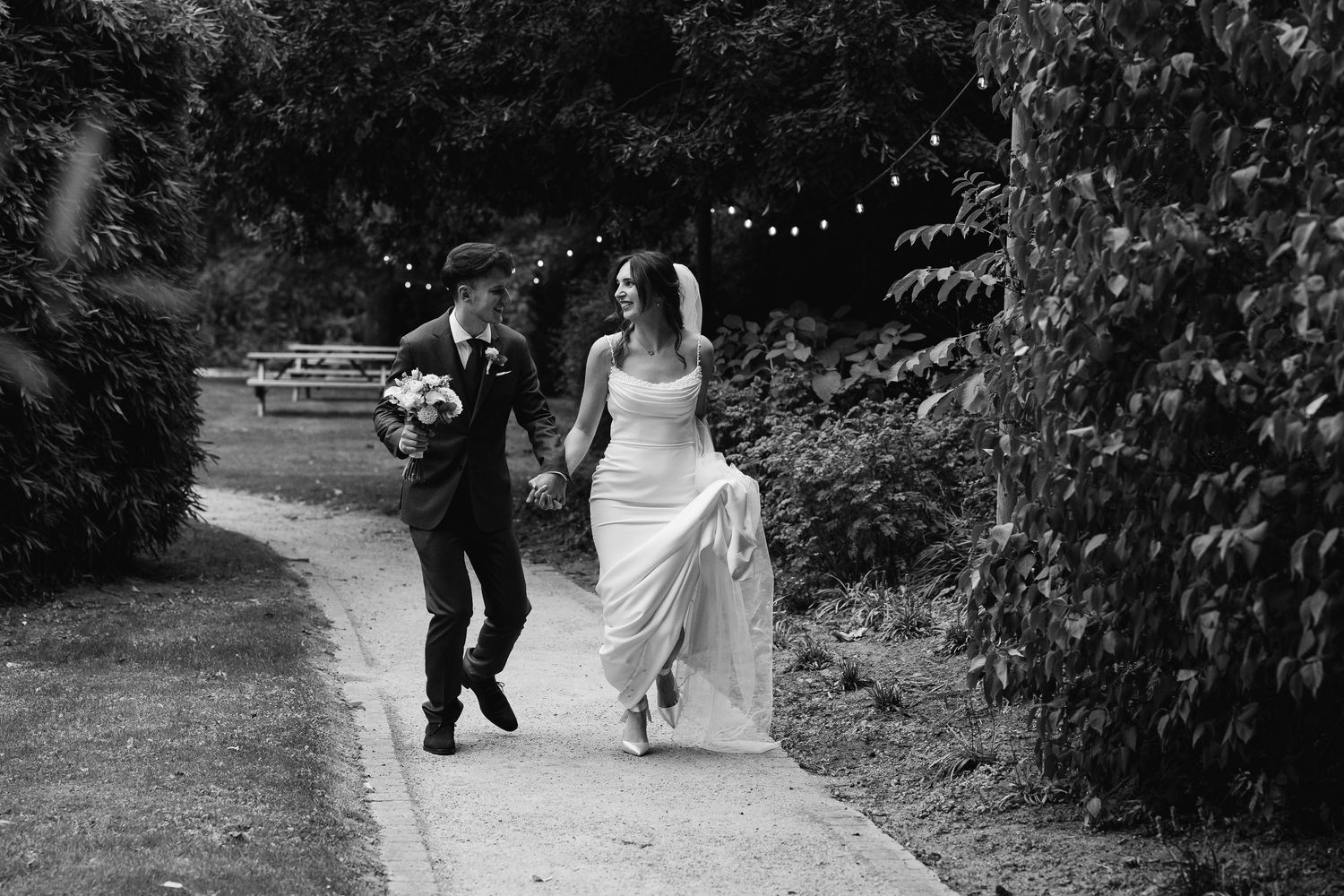 Black and white photo of a bride and groom holding hands and running along a garden path on their wedding day.