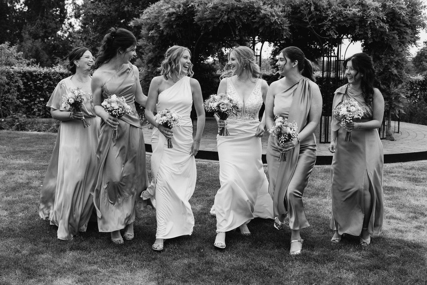 Black and white photo of bride and five bridesmaids walking together laughing in garden holding bouquets.
