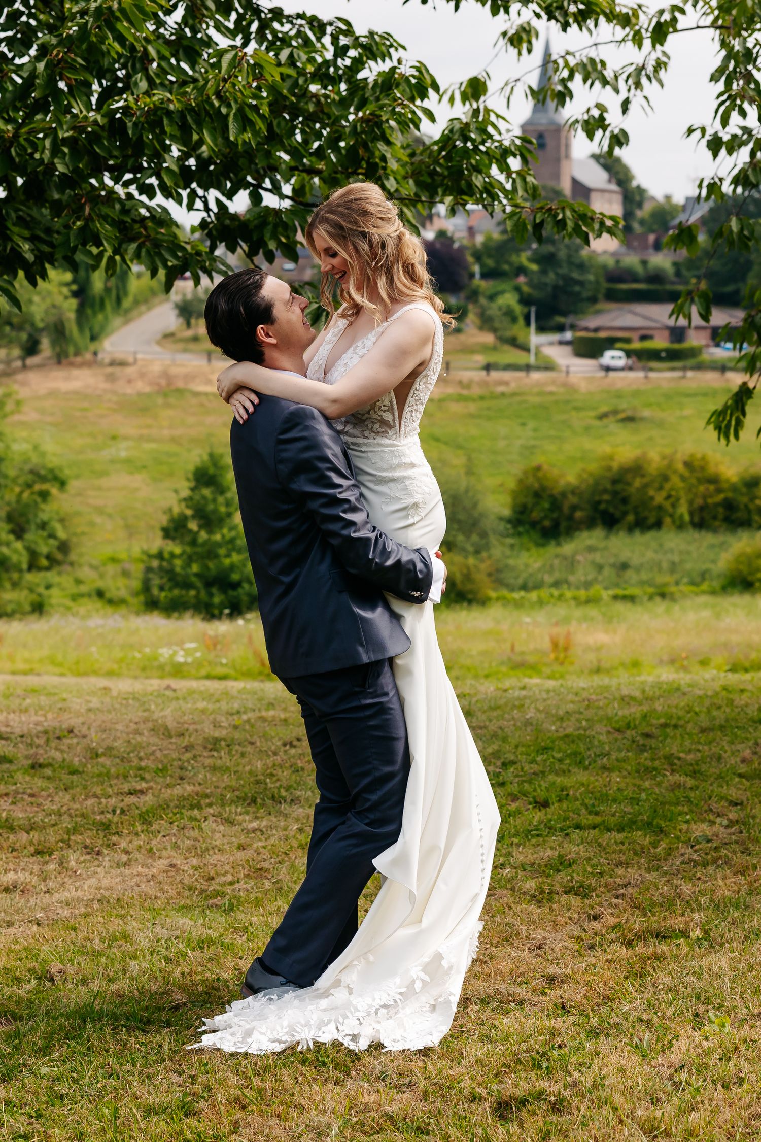 Groom lifting bride in white lace wedding dress outdoors on green grass with lush countryside background.