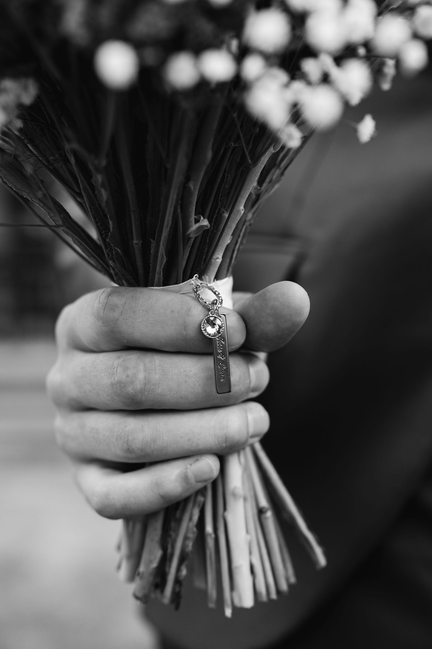 Black and white close-up of groom holding wedding bouquet with decorative charm on stems.