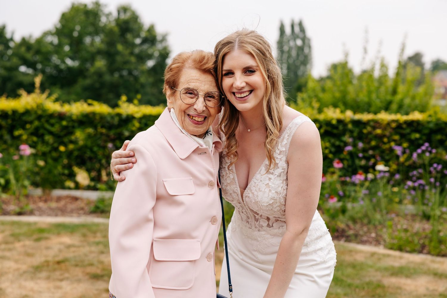 A radiant bride in a lace wedding dress smiles alongside an elderly woman in a pink blazer outdoors.