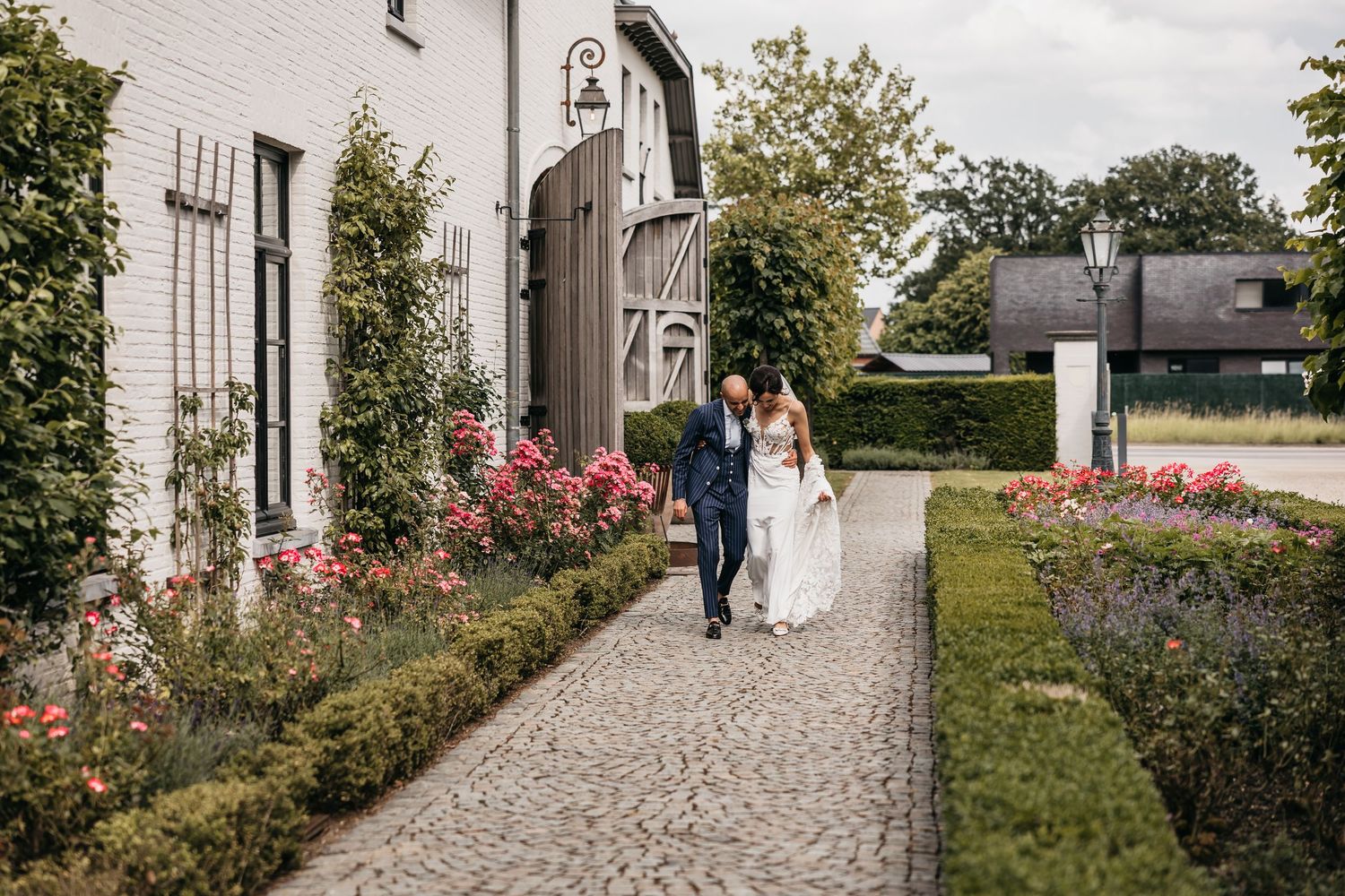 Bride and groom walk hand-in-hand along a cobblestone path beside a white building with rose gardens.