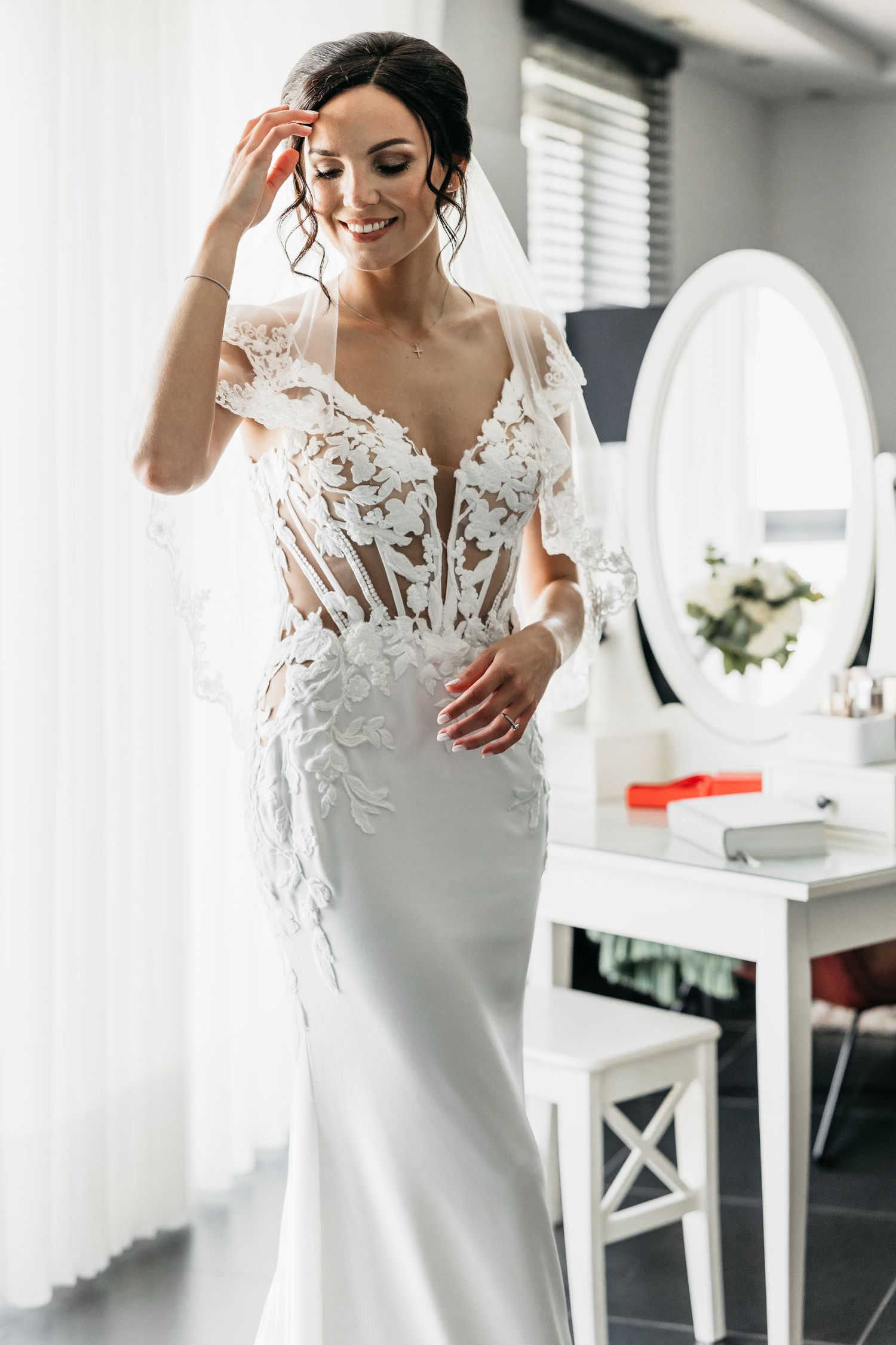 Beautiful smiling bride in intricate lace wedding dress adjusting veil near white dressing table.