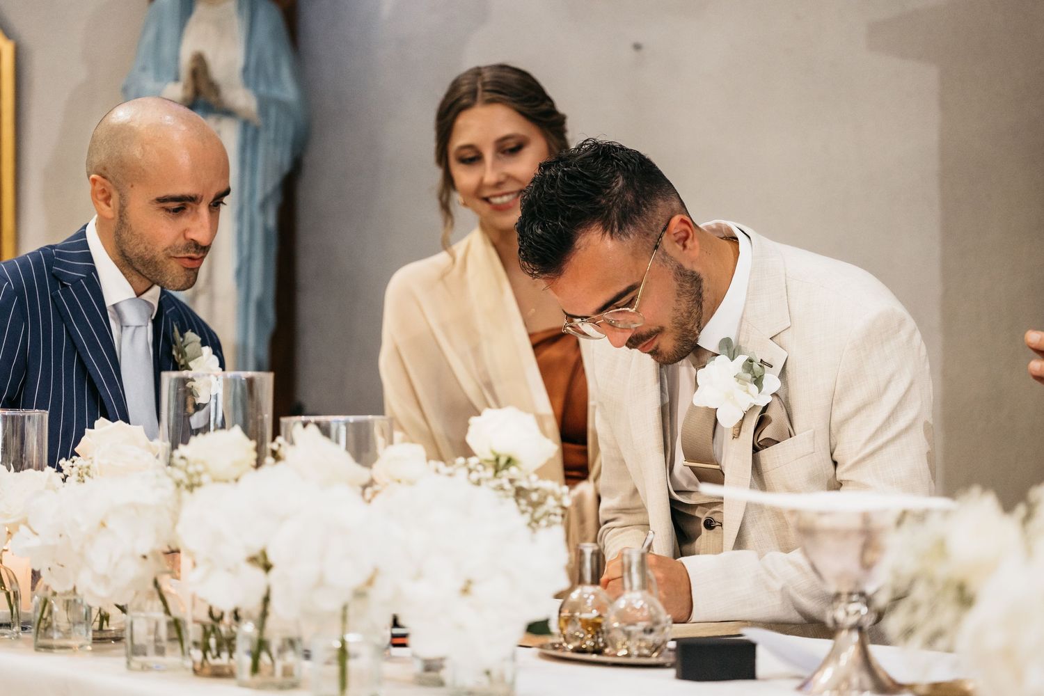 Groom in cream suit signing wedding register as bride and guest look on at floral-decorated altar.
